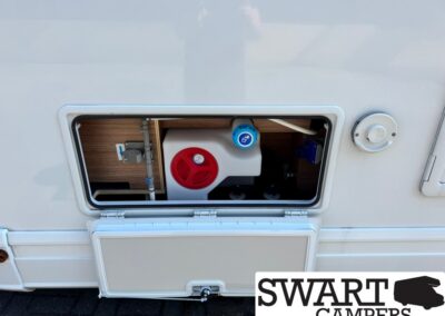 Open external service panel of a white camper showing plumbing valves: red circular valve and blue fill cap visible inside the compartment