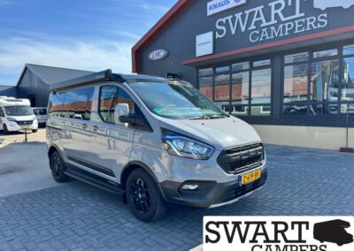 Front three-quarter view of a silver camper van with black wheels and roof rack, parked outside Swart Campers showroom.