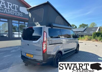 Gray Ford Nugget Trail camper van with a raised pop-up roof parked in a lot beside a Swart Campers building showing large signage in the background.