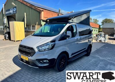 White Ford camper van with a raised pop-top roof parked on a brick lot; SWART CAMPERS logo visible in the corner.