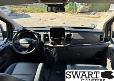 Interior view of a Ford camper van cockpit: steering wheel, gear shifter, center touchscreen, and front seats, with a branding watermark 'SWART CAMPERS' in the lower-right corner.
