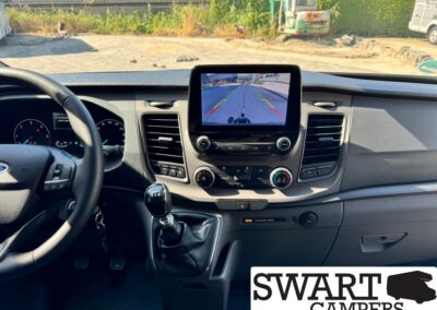 Interior of a van cockpit with a center touchscreen showing a rear-view camera feed and parking guides, gear shift, and climate controls nearby.