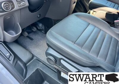 Interior view of a vehicle cabin showing the driver's seat with a manual height/adjustment knob, pedals, a cup holder, and floor mat; Swart Campers watermark present