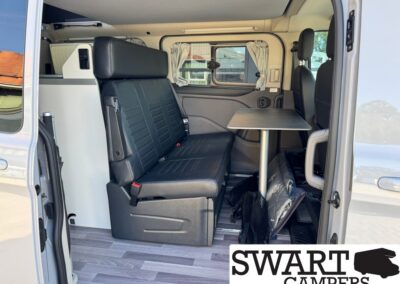 Interior of a camper van: black leather bench seat with a fold-down table, windows with curtains, and a light wood floorSA