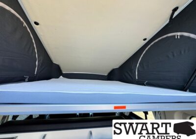 Interior of a pop-up camper: raised blue mattress on a bed platform with dark fabric tent walls at the sides and rear