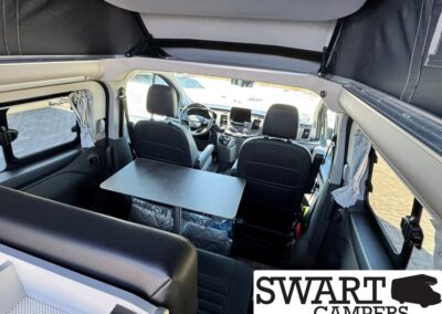 Interior of a camper van with a rectangular table between the front seats and a view of the driver’s area; side windows with curtains visible, Swart Campers logo in corner.