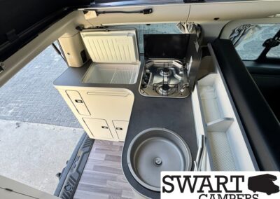 Compact camper van kitchenette with sink, two-burner stove, and small fridge with storage cabinets nearby.