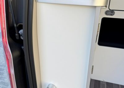 Built-in utility cabinet in a camper van with a wrapped coiled hose on a metal top, beside the doorway.