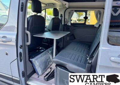Interior of a camper van: back seating with a metal pedestal table between the seats and gray upholstery, windows with curtains on the side.