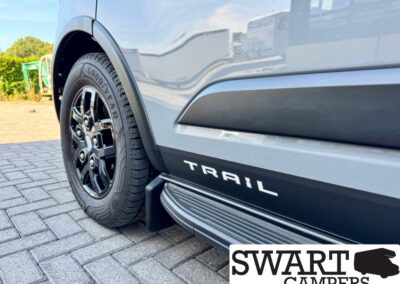 Close-up of a gray camper van side with a Goodyear tire, black wheel arch trim, and the word TRAIL along the lower panel; SWART CAMPERS logo visible in the corner.
