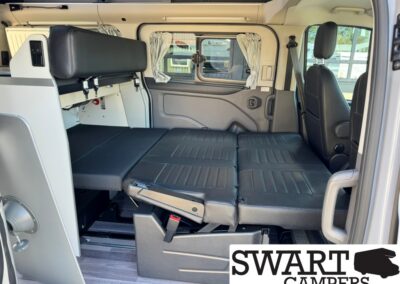 Interior of a camper van with a gray fold-out bed spanning width, cushions in bed mode, driver seat at right, side window with a curtain, compact storage panel on left.