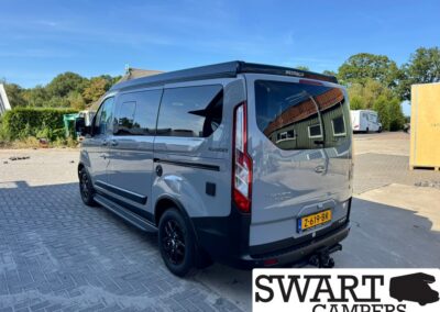 Grey Ford Transit Nugget camper van parked in a paved lot, rear and passenger side visible, with a Westfalia roof rack and black wheels; license plate 7-619-BR, Swart Campers watermark in corner.