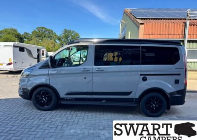 Side view of a gray camper van with black wheels and tinted windows parked on a brick lot, Solar panels on the roof and Swart Campers watermark bottom right.