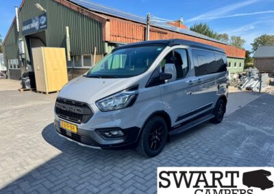 Gray Ford Transit camper van with black wheels parked outside a workshop; SWART CAMPERS logo visible in the corner.