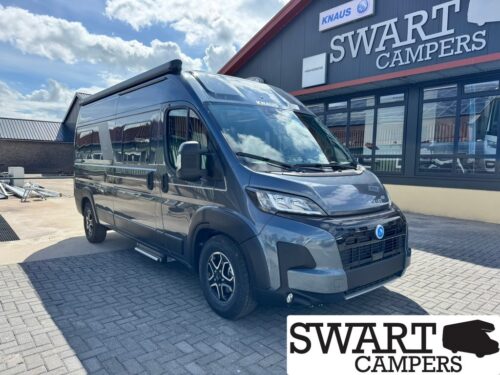 Front three-quarter view of a dark gray camper van with a high roof parked outside a Swart Campers storefront with a bold sign above the windows.