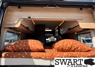 Camper van rear interior with a bed covered in orange polka-dot bedding and two pillows, SWART CAMPERS logo in the corner.