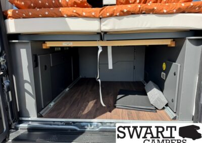 Camper van rear interior with a raised bed and orange polka-dot bedding; storage space beneath and wood-look flooring