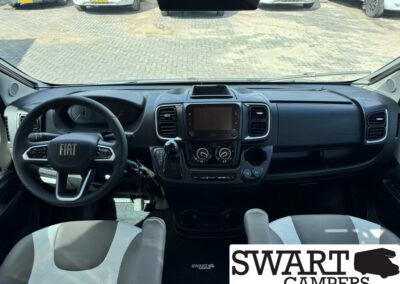 Interior view of a Fiat camper van cockpit showing the steering wheel, central touchscreen, climate controls, gear shifter, and front seats; Swart Campers logo in the corner.
