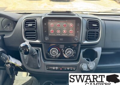 Camper van dashboard with central touchscreen, climate dials, gear shifter, cup holder, and Swart Campers logo watermark in corner.