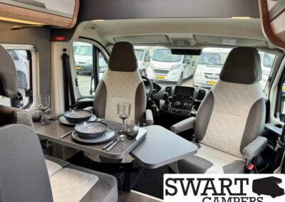Interior of a camper van with a folding table set for a meal: plates, bowls, glasses, and cutlery on a gray table; two front seats with light upholstery; dashboard visible through the windshield; SWART CAMPERS logo in the corner.