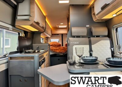 Interior of a compact camper van with a dining table set for two, kitchenette on left, and a bed at the rear.