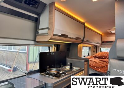 Interior of a modern camper van: compact kitchen with a two-burner stove and sink, overhead storage cabinets with warm LED lighting, and a bed with orange polka-dot bedding visible in the rear.