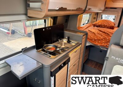 Interior of a camper van showing a compact kitchenette with two-burner stove, frying pan, and sink, adjacent counter space, and a bed with an orange polka-dot blanket in the back. SWART CAMPERS logo visible in the corner.