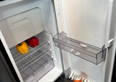 Open compact RV fridge with yellow and red bell peppers on the lower rack and a can of Fanta on the door shelf; Swart Campers watermark present.