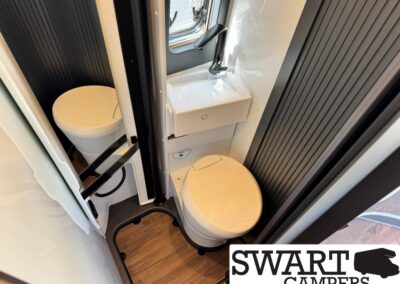 Compact camper bathroom with a small toilet, wall-mounted sink, and mirror in a narrow room with wood-look floor and dark vertical panels.