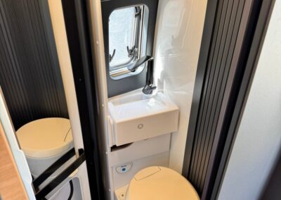 Compact camper bathroom with a small sink, faucet, window, and a toilet; hairbrush rests on a shelf above.