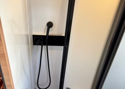 Compact camper shower: handheld shower on hose, white walls, black shelf, and sliding frosted door.