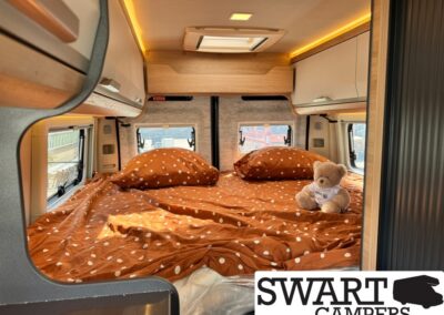 Bed area inside a camper van with orange polka-dot bedding, two pillows, and a small teddy bear against the back wall.
