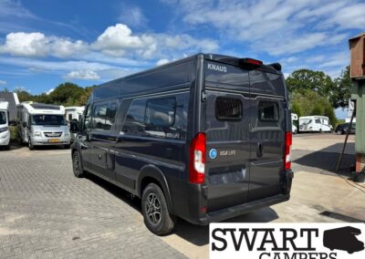 Rear and side view of a dark blue Knaus Box Life camper van parked in a lot with other RVs under a blue sky on a sunny day (Swart Campers logo in corner).