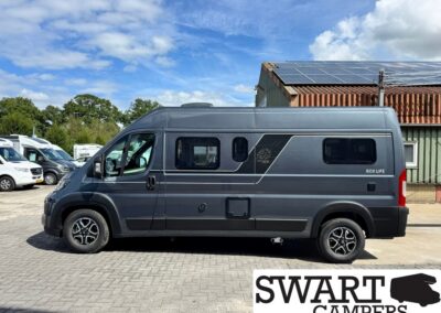 Dark gray camper van with solar panels on the roof, parked in a lot beside other white vans; SWART CAMPERS watermark bottom right.