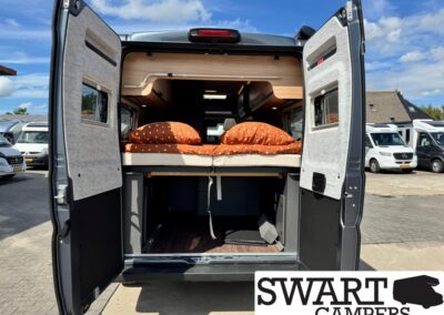 Rear view of a camper van with a raised bed and wood storage, open doors, showing a compact living area under clear blue sky.