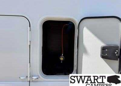 Open storage compartment on a camper van showing an orange hose and gauge inside; SWART CAMPERS watermark bottom-right.