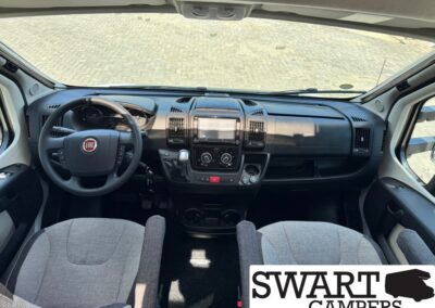 Interior view of a Fiat camper van cockpit: steering wheel, central touchscreen, climate controls, and front seats seen from the passenger side