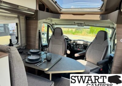 Camper van interior with a dining table set for two, two front captain chairs, plates and glasses, and a skylight overhead (Swart Campers branding visible).