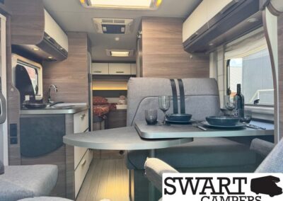 Interior of a modern camper van with a dining table set for two, grey seats, kitchenette, and a bed area at the rear, plus a SWART CAMPERS logo in the corner.