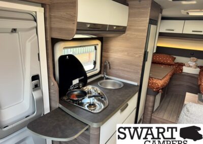 Compact camper kitchen with a two-burner stove, sink, and small fold-out counter next to a window. A bed area with polka-dot bedding is visible in the background.