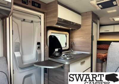 Interior of a compact camper kitchen: fold-out table, two-burner stove with cover, sink, and wood-tone cabinetry beside a window.