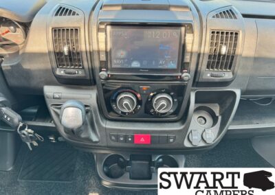 Center dashboard of a van: touchscreen infotainment system, two climate dials, gear lever to the left, USB/12V ports on the right. SWART CAMPERS watermark bottom-right.