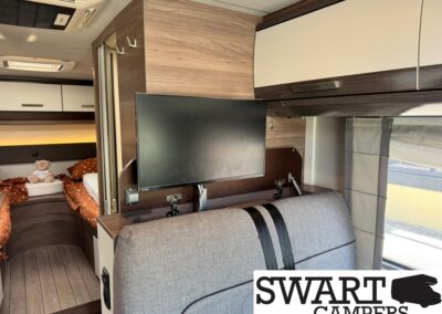 Interior of a camper van showing a wall-mounted flat-screen TV above a gray sofa, with a bedroom area featuring orange bedding and a teddy bear in the back.