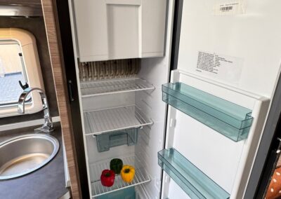Open camper fridge with two bell peppers (red and yellow) on the lower shelf and a small blue basket at the bottom; door shelves are mostly empty with a can on the door.
