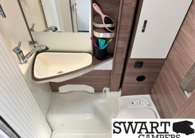 Compact caravan bathroom with a white sink, chrome faucet, mirrored wall, and woodgrain storage panel.