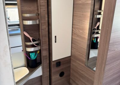 Compact camper bathroom with wood panels, a tall storage cabinet, mirrors, and open shelves holding a brush and toiletries.