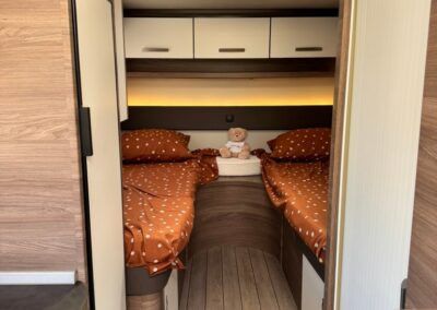 Camper bedroom with two single beds, orange polka-dot bedding, a teddy bear between them, and white overhead cabinets.
