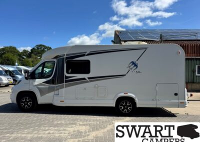 Side view of a white motorhome with black and gray graphics, parked on a paved lot under a blue sky with a building and solar panels nearby.