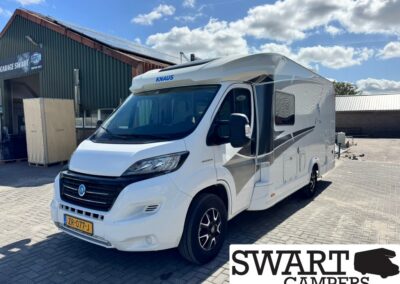 Front-side view of a white Knaus motorhome with gray graphic stripes parked on a paved lot beside a workshop; Dutch license plate visible (ZR-077-J).