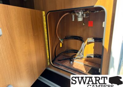 Open camper cabinet showing propane hose, valve, and regulator with storage rails and straps inside a wood-paneled compartment.